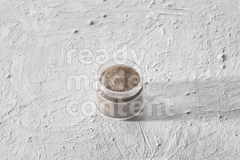 A glass jar full of black pepper powder on a textured white flooring