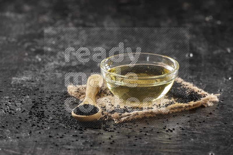 A glass bowl full of black seeds oil and wooden spoon full of black seeds with seeds spreaded on burlap fabric on a textured black flooring