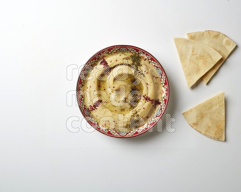 Hummus in a red plate with patterns garnished with zaatar & sumak on a white background