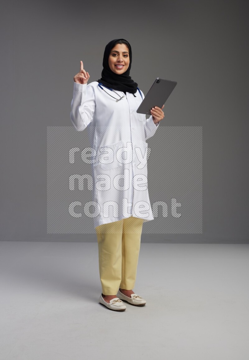 Saudi woman wearing lab coat with stethoscope standing working on tablet on Gray background