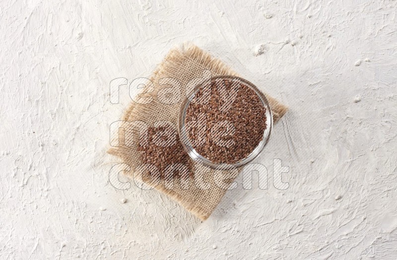 A glass bowl full of flax seeds and a bunch of seeds on burlap fabric on a textured white flooring