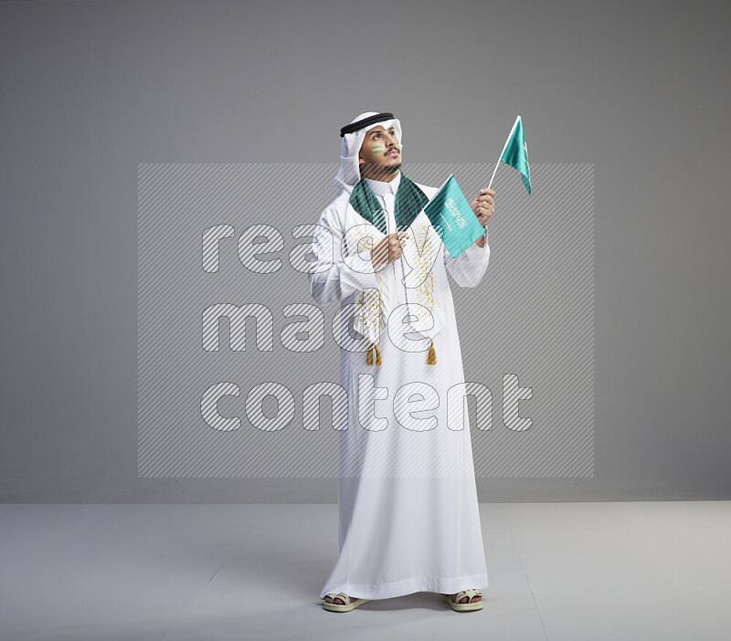 A saudi man standing wearing thob and white shomag with face painting and saudi flag scarf and holding small saudi flag on gray background