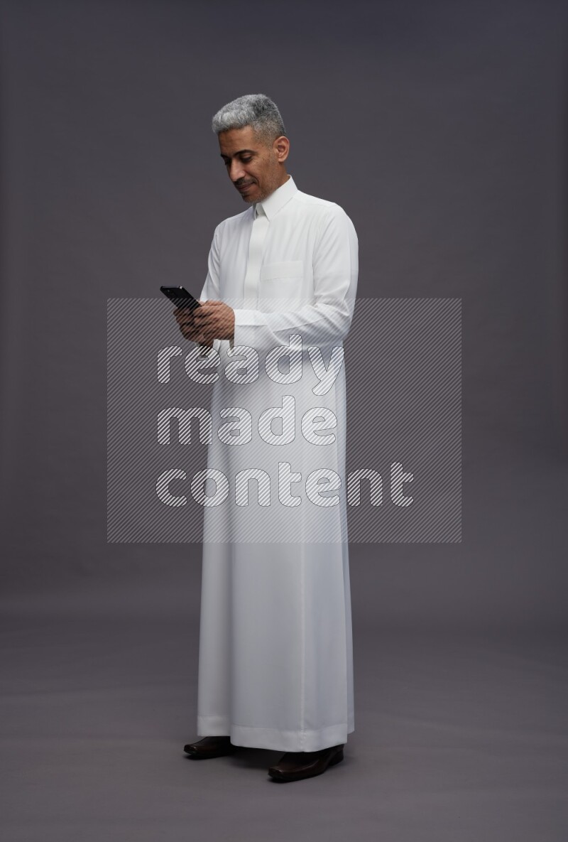 Saudi man wearing thob standing texting on phone on gray background