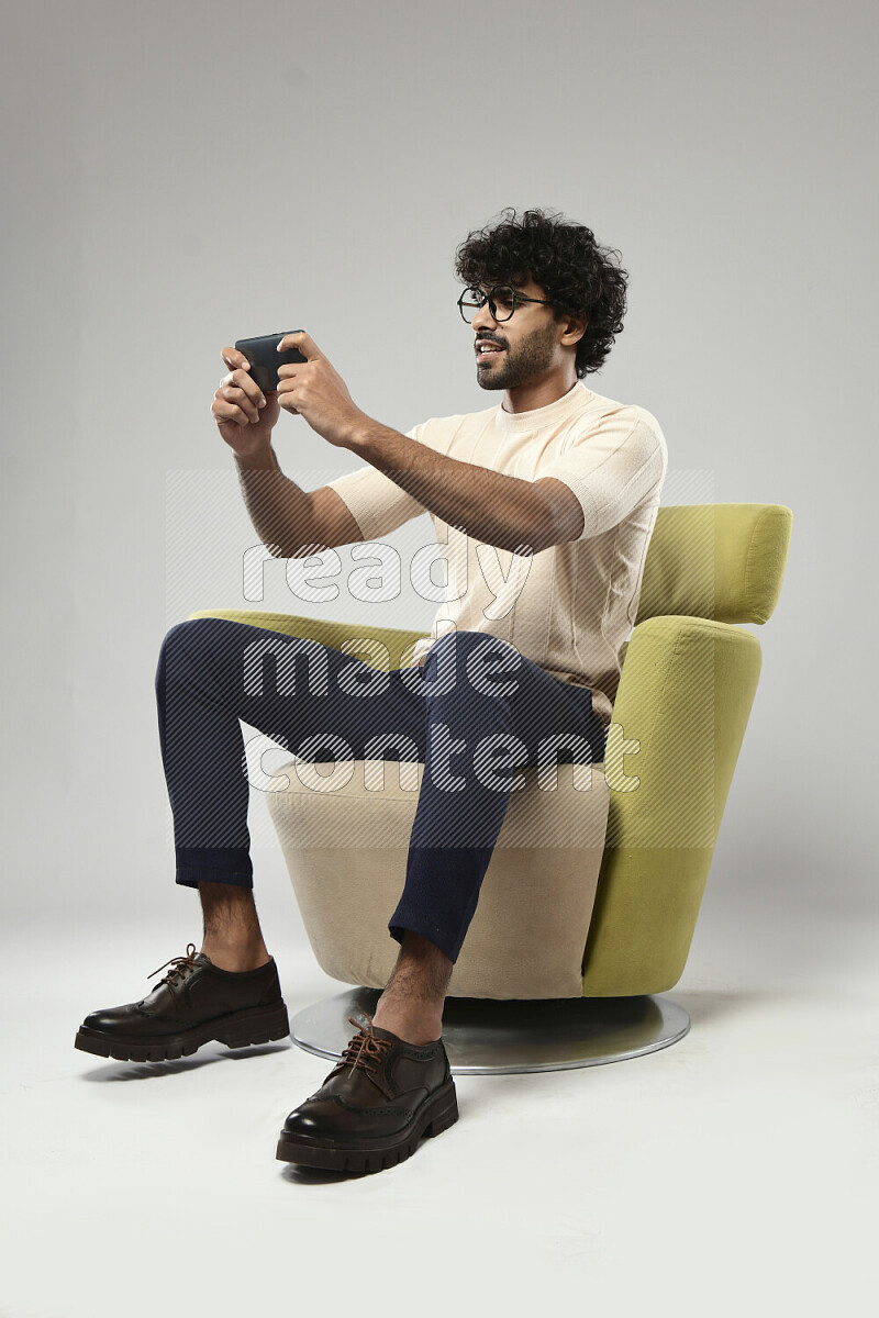 A man wearing casual sitting on a chair gaming on the phone on white background