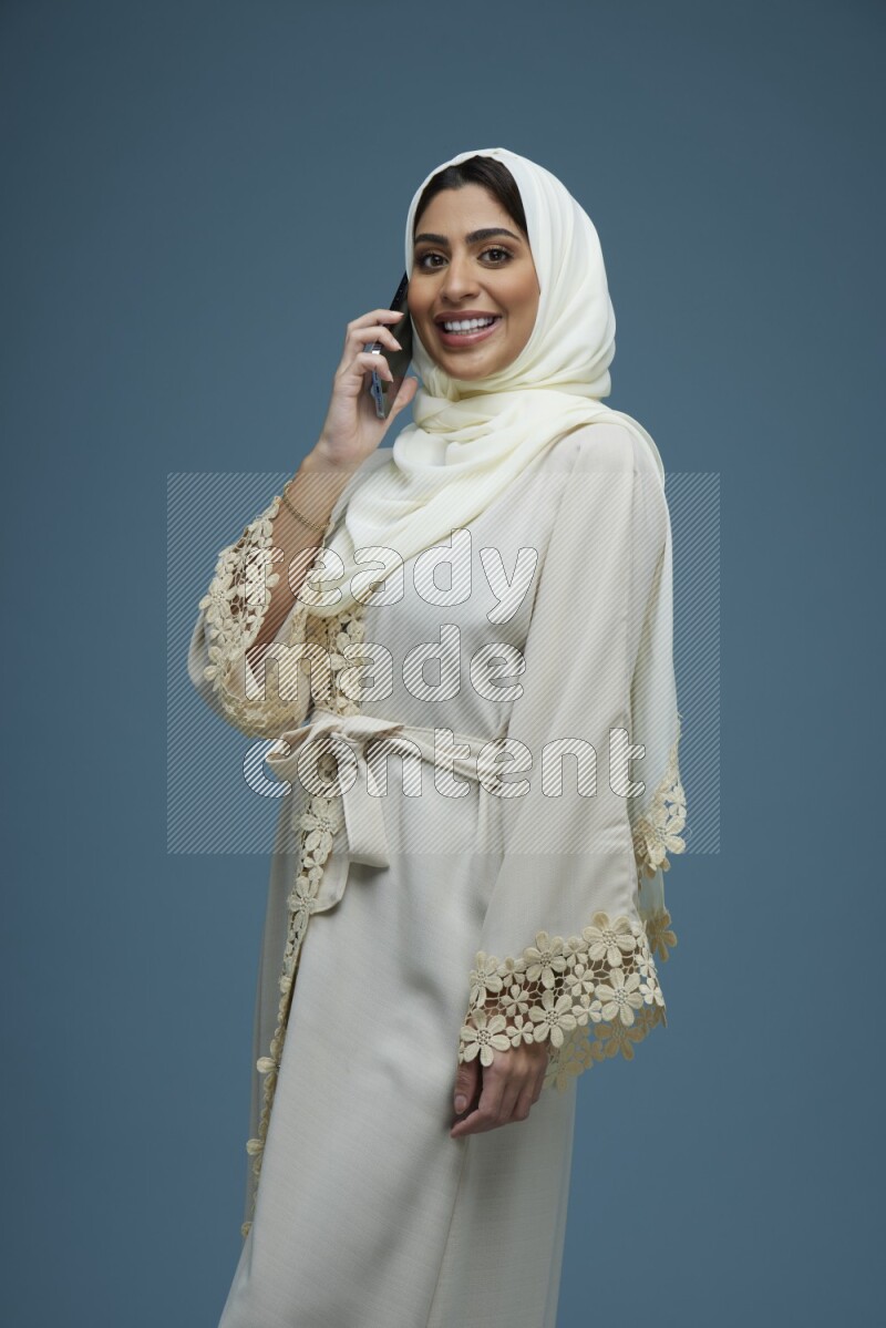 A Saudi woman having a call in a blue background wearing an off-white Abaya Hijab