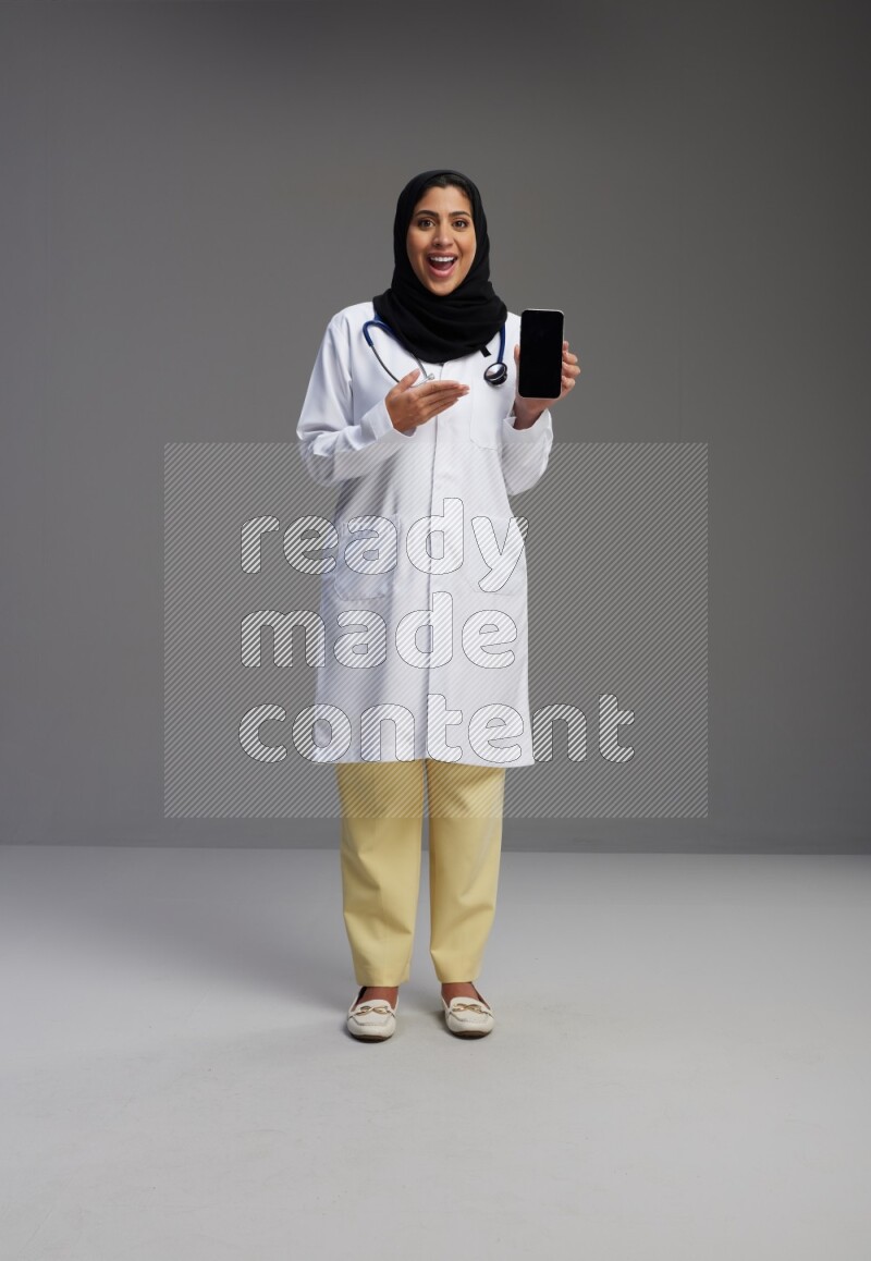 Saudi woman wearing lab coat with stethoscope standing showing phone to camera with sign in the back on Gray background