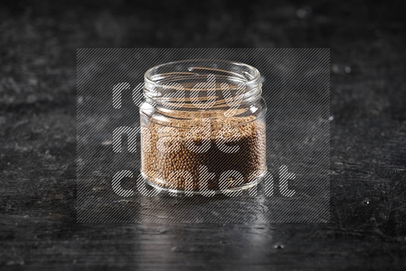 A glass jar full of mustard seeds on a textured black flooring