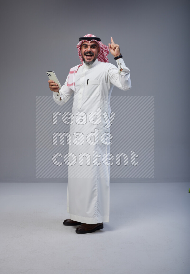 Saudi man Wearing Thob and red Shomag standing texting on phone on Gray background