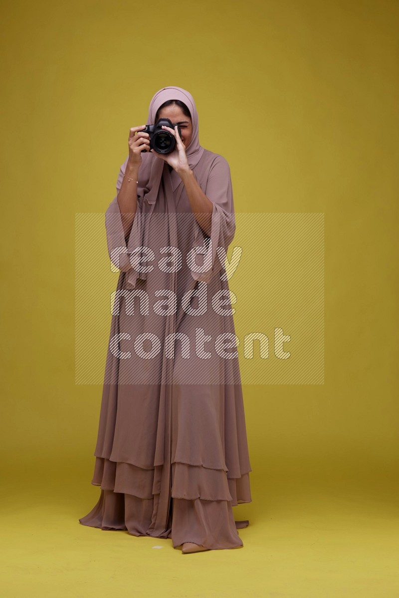 A woman Holding a Camera on a Yellow Background wearing Brown Abaya with Hijab