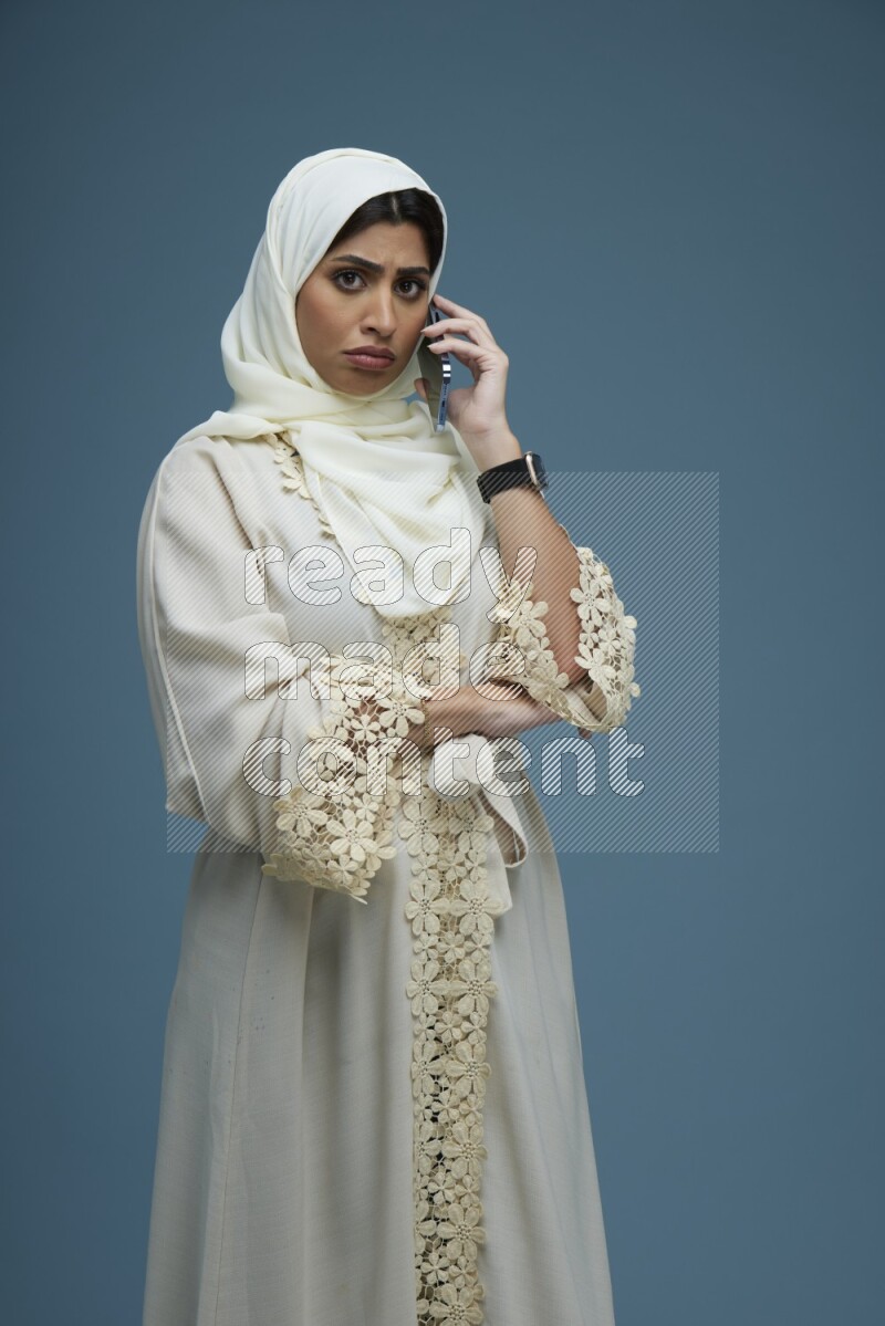 A Saudi woman having a call in a blue background wearing an off-white Abaya Hijab