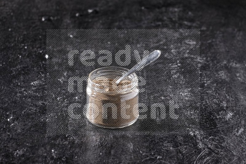 بودرة الفلفل الأسود في وعاء زجاجي مع ملعقة معدنية علي خلفية سوداء