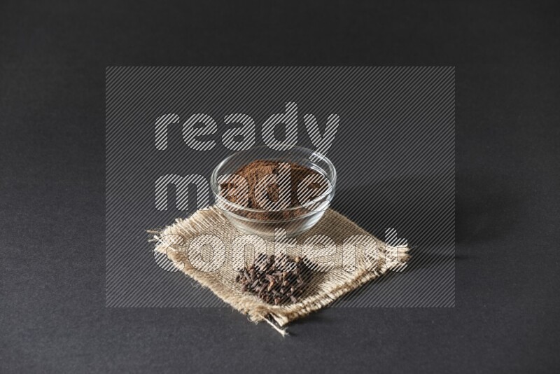 A glass bowl full of cloves powder with cloves grains on a burlap piece on a black flooring