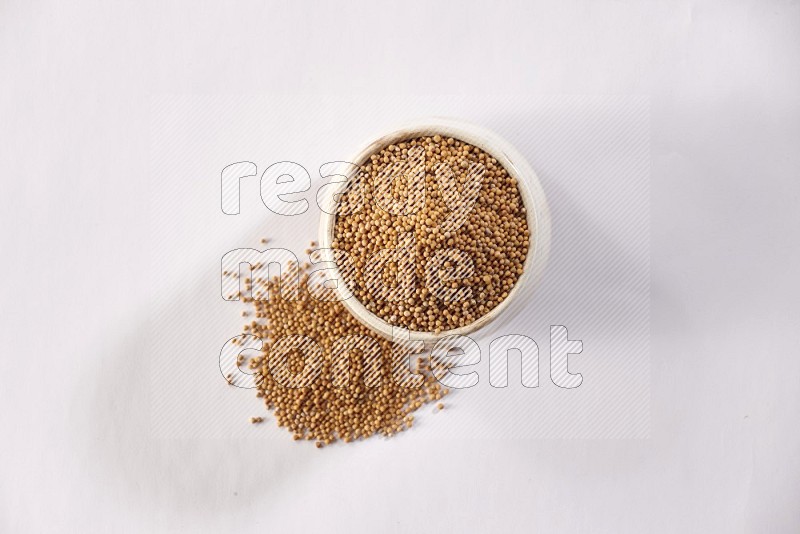 A beige pottery bowl full of mustard seeds and more seeds spread on a white flooring