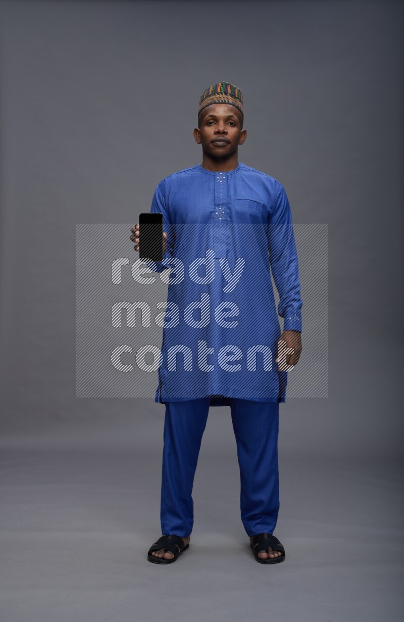 Man wearing Nigerian outfit standing showing phone to camera on gray background