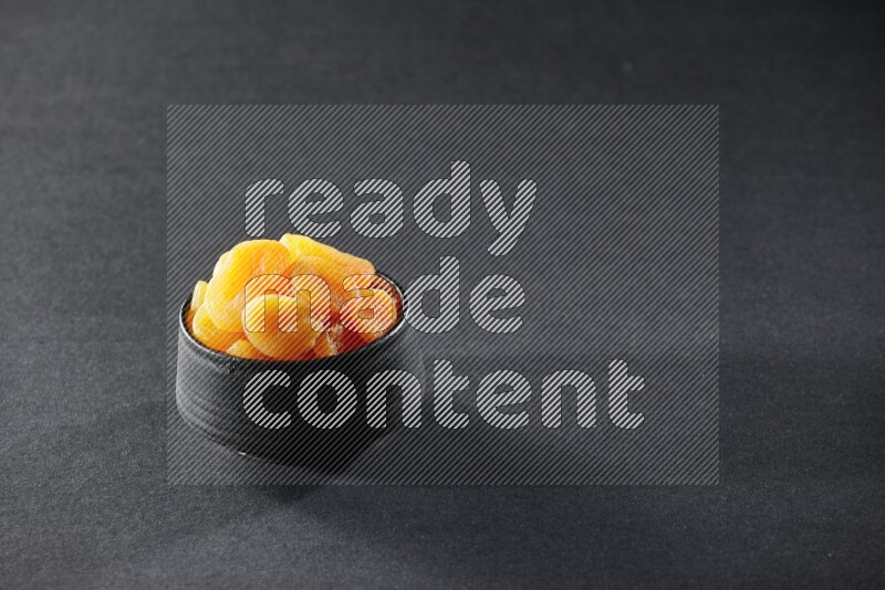 A black pottery bowl full of dried apricots on a black background in different angles