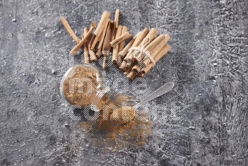 Herbal glass jar full cinnamon powder flipped and a metal spoon full of powder, cinnamon sticks stacked and bounded in the back on textured black background in different angles