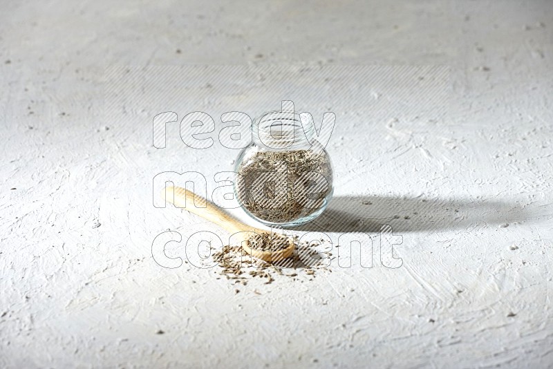 A glass spice jar and wooden spoon full of cumin seeds on textured white flooring