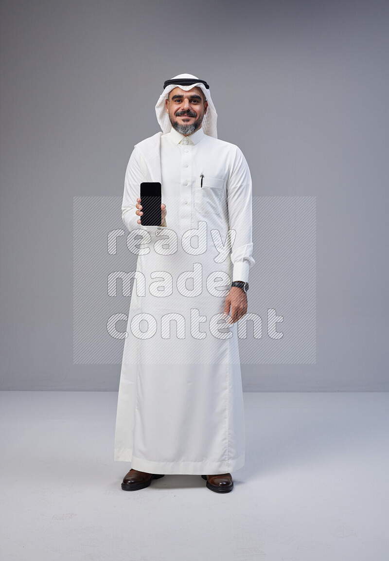 Saudi man Wearing Thob and white Shomag standing showing phone to camera on Gray background