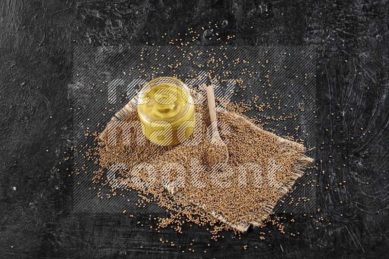 A glass jar full of mustard paste set on a burlap piece and a wooden spoon full of mustard seeds on a textured black flooring in different angles