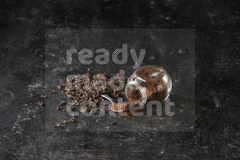 A flipped glass spice jar and a metal spoon full of cloves powder with cloves spread on textured black flooring