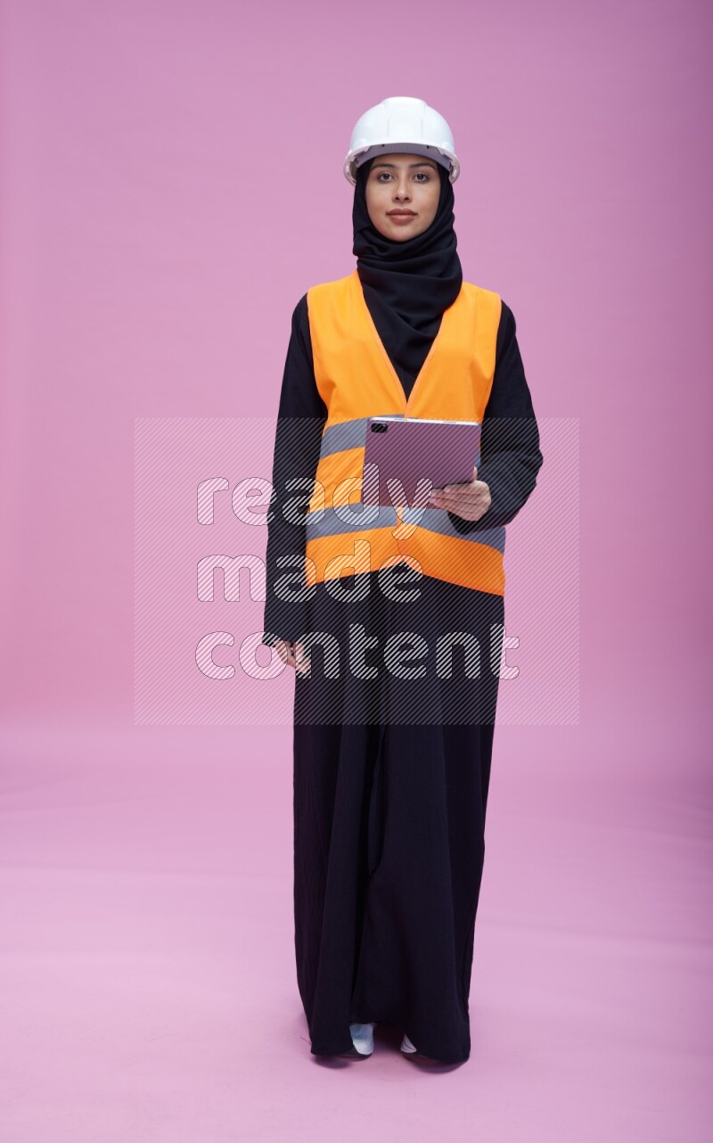 Saudi woman wearing Abaya with engineer vest and helmet standing working on tablet on pink background