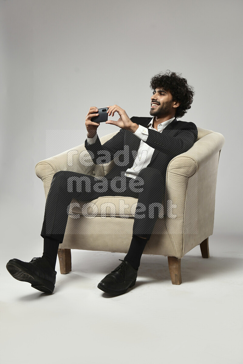رجل يرتدي ملابس رسمية يجلس علي كرسي و يصور بهاتفه علي خلفية بيضاء