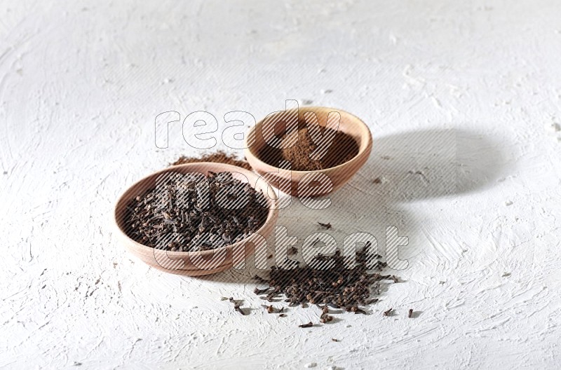 2 wooden bowls full of cloves powder and whole cloves on a textured white flooring