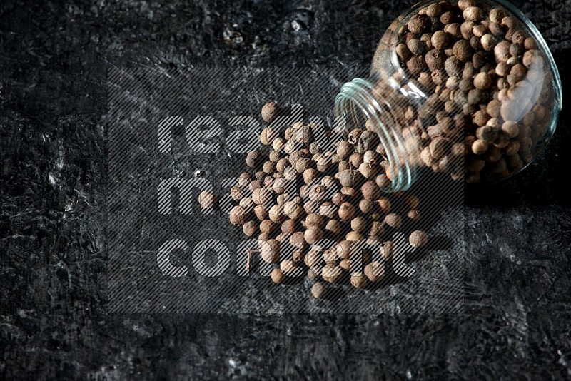 A flipped glass spice jar full of allspice whole balls and the balls spilled out of it on a textured black flooring