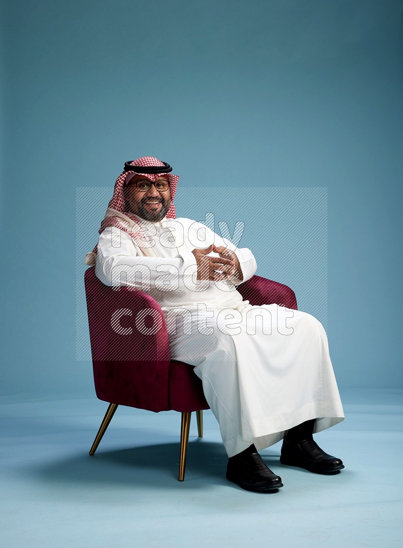 Saudi Man with shimag sitting on chair Interacting with the camera on blue background