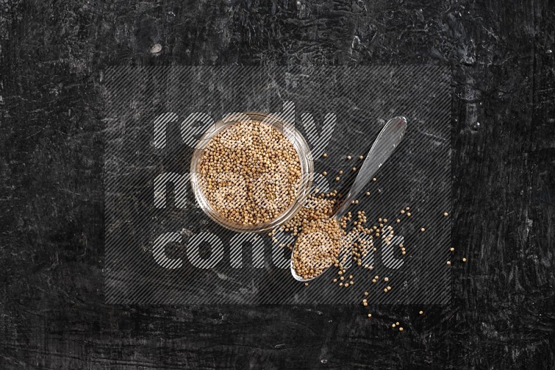 A glass jar and a metal spoon full of mustard seeds on a textured black flooring