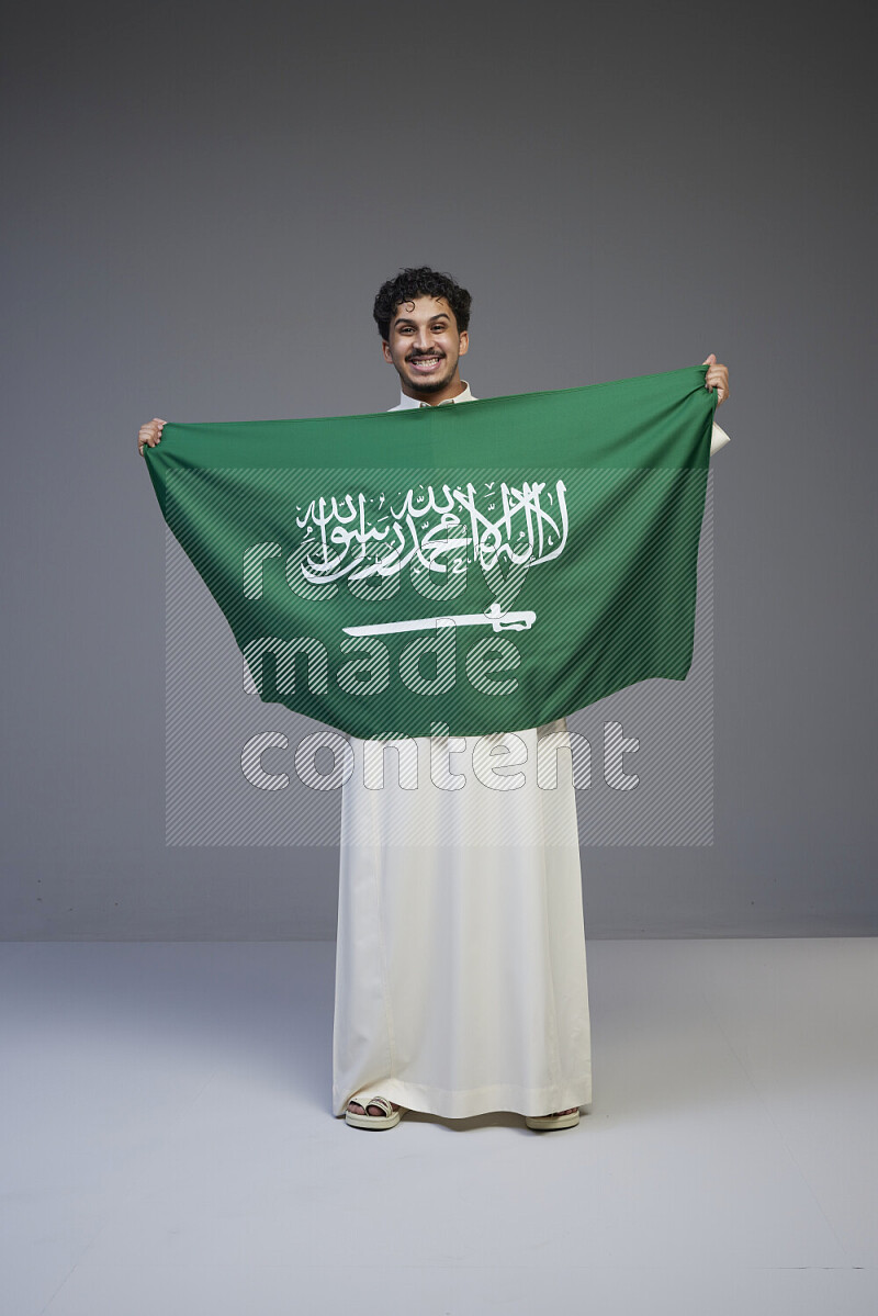 A Saudi man standing wearing thob holding big Saudi flag on gray background