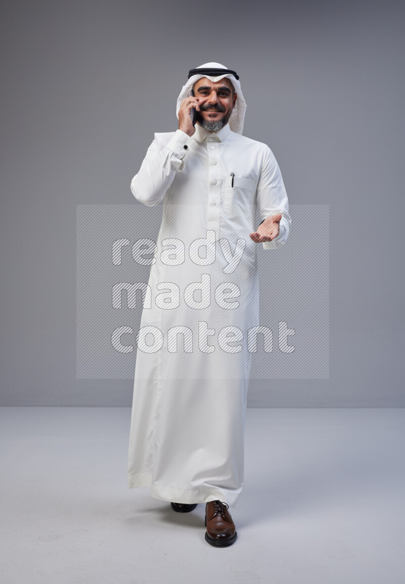 Saudi man Wearing Thob and white Shomag standing talking on phone on Gray background