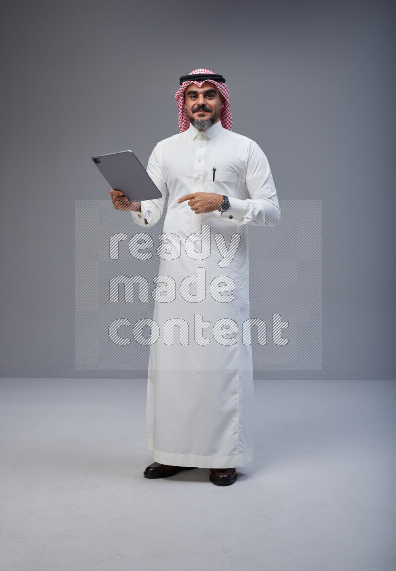 Saudi man Wearing Thob and red Shomag standing working on tablet on Gray background