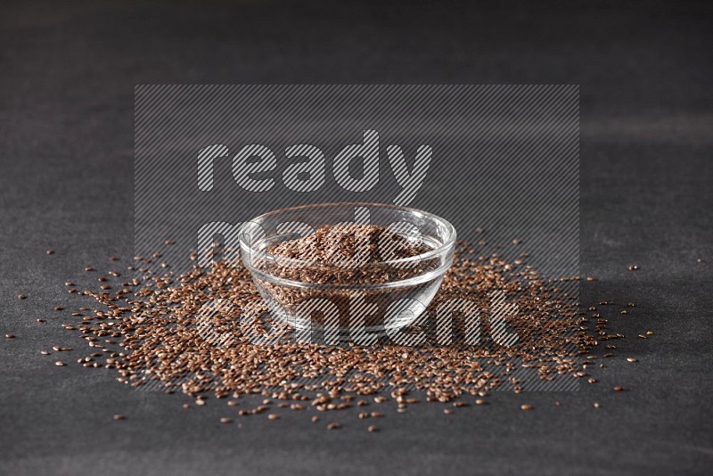 A glass bowl full of flaxseeds surrounded by the seeds on a black flooring