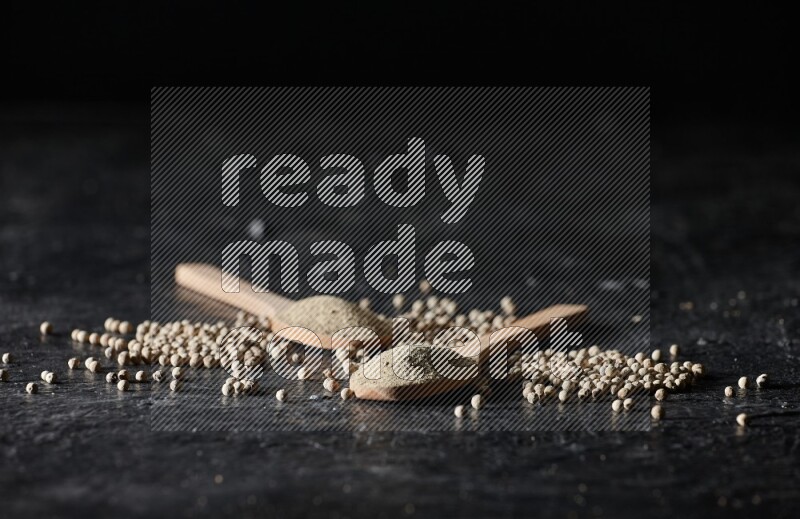 2 wooden spoons full of white pepper powder with white pepper beads on textured black flooring
