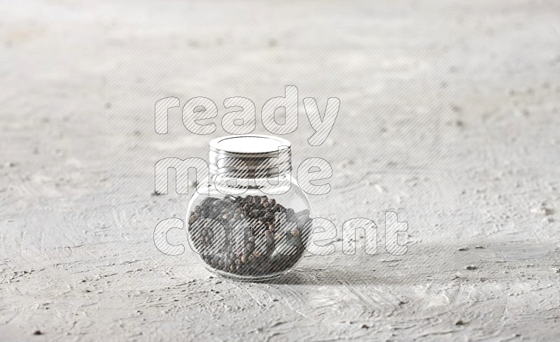 A glass spice jar full of black pepper on textured white flooring