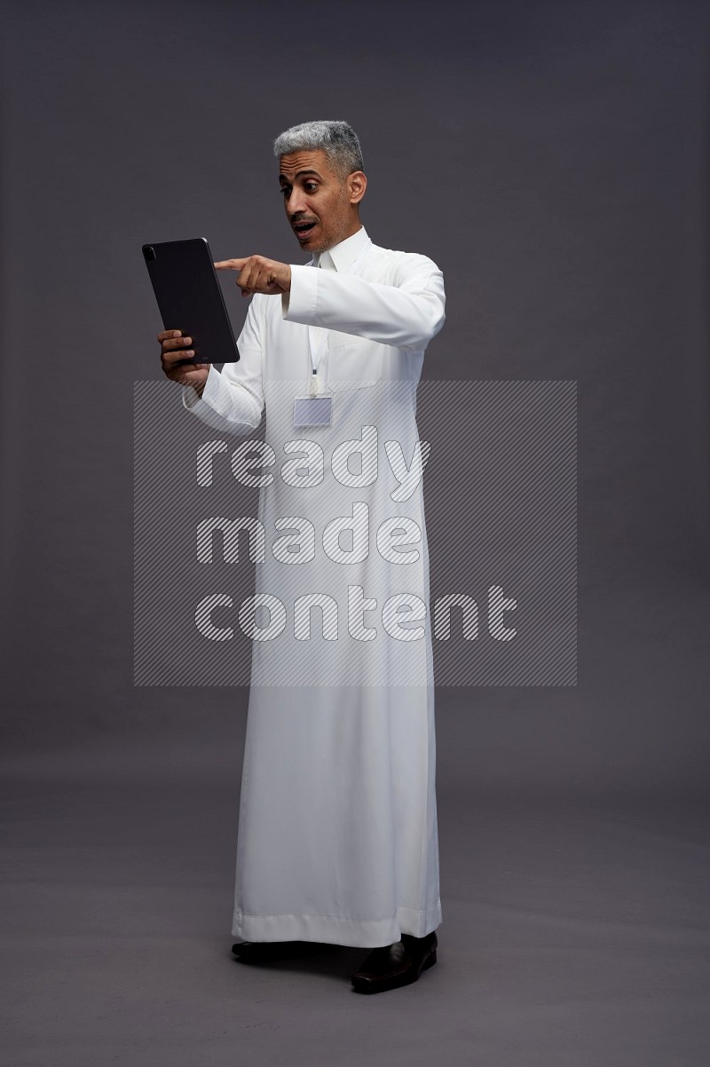 Saudi man wearing thob with neck strap employee badge standing working on tablet on gray background