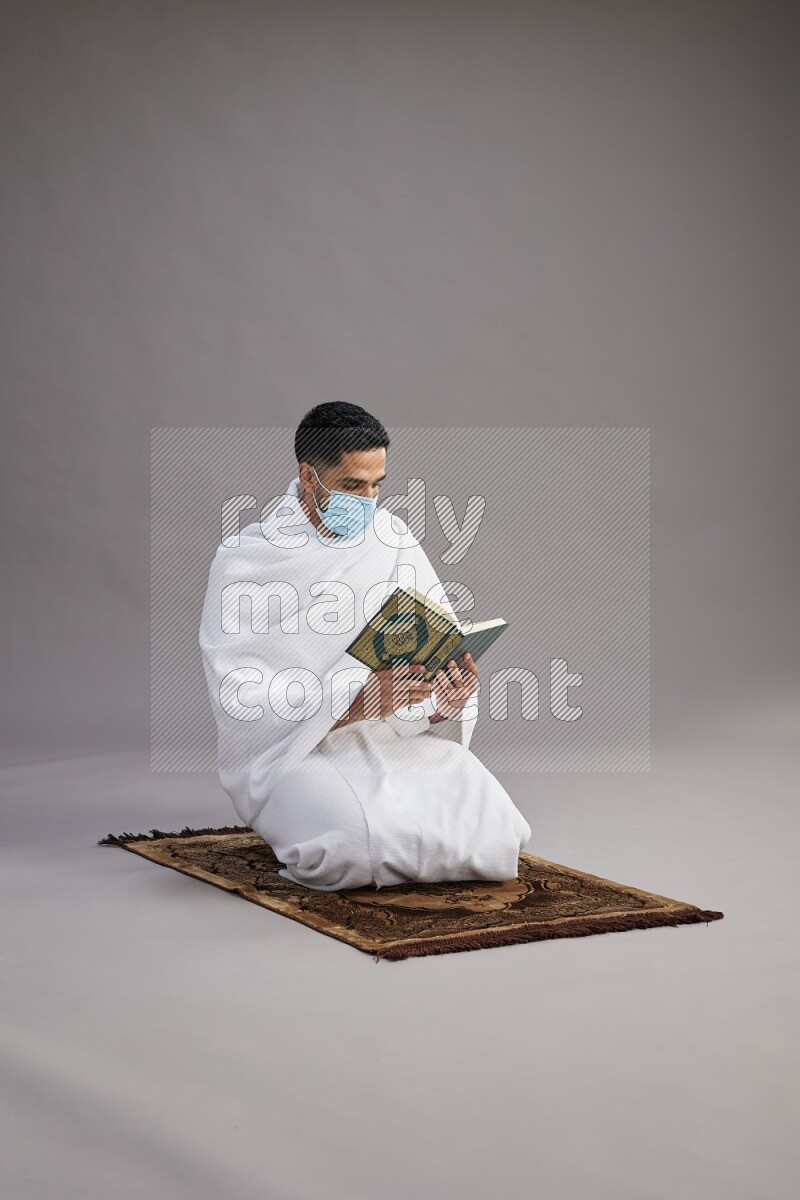 A man wearing Ehram with face mask sitting on floor reading quran on gray background