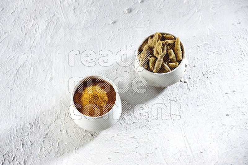 2 beige pottery bowl full of turmeric powder and dried turmeric whole fingers on textured white flooring