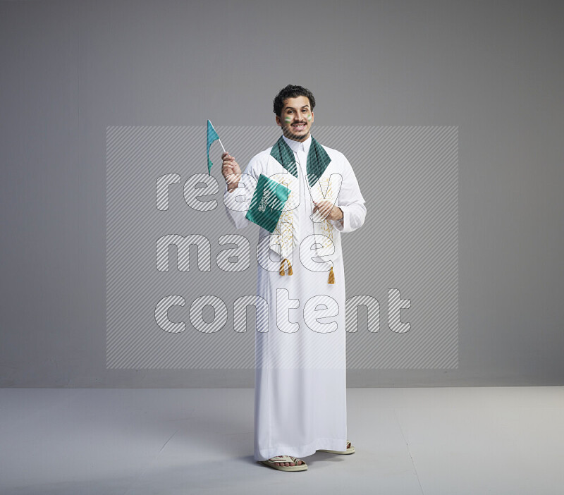 A Saudi man standing wearing thob and saudi flag scarf with face painting holding small Saudi flag on gray background