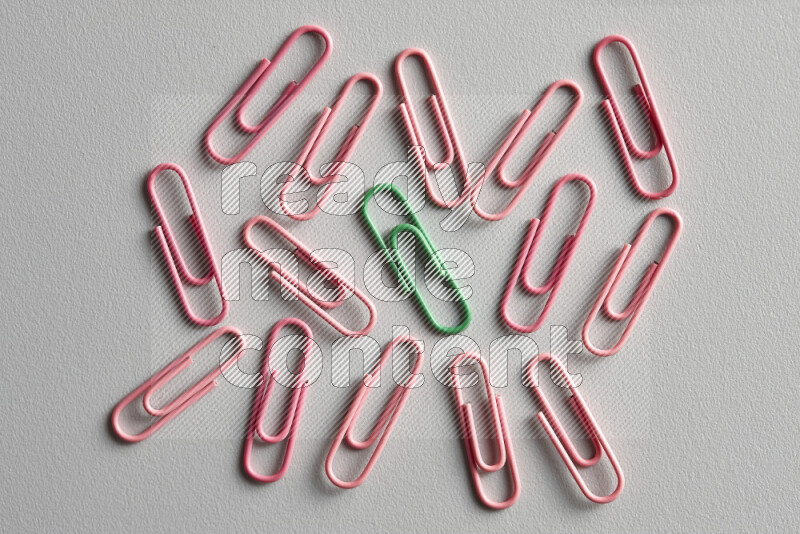 A green paperclip surrounded by bunch of pink paperclips on grey background