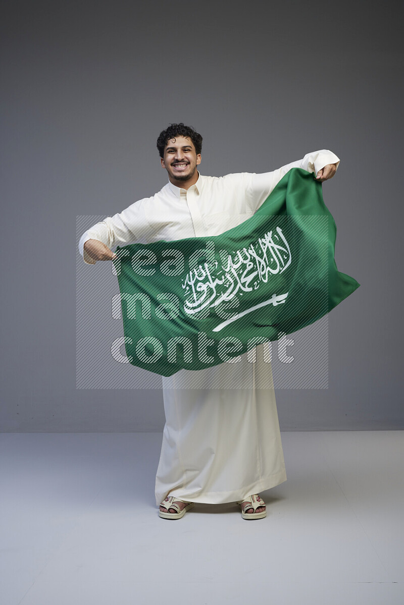 A Saudi man standing wearing thob holding big Saudi flag on gray background