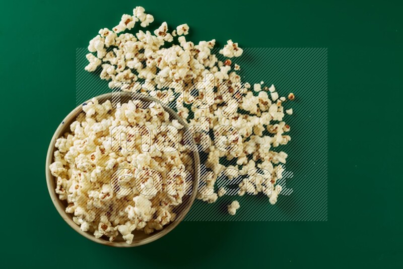 A beige ceramic bowl full of popcorn with popcorn beside it on a green background in a top view shot