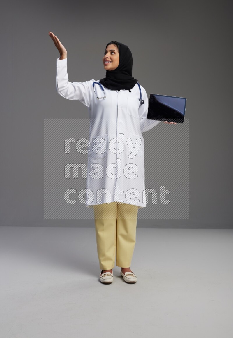 Saudi woman wearing lab coat with stethoscope standing showing tablet to camera with sign in the back on Gray background