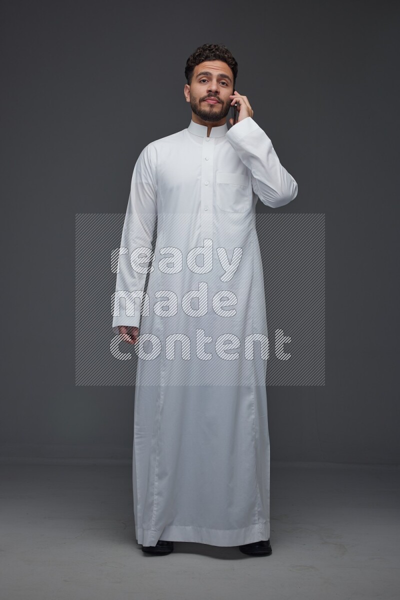 A Saudi man wearing Thobe and talking in the phone while standing and making different poses eye level on a gray background