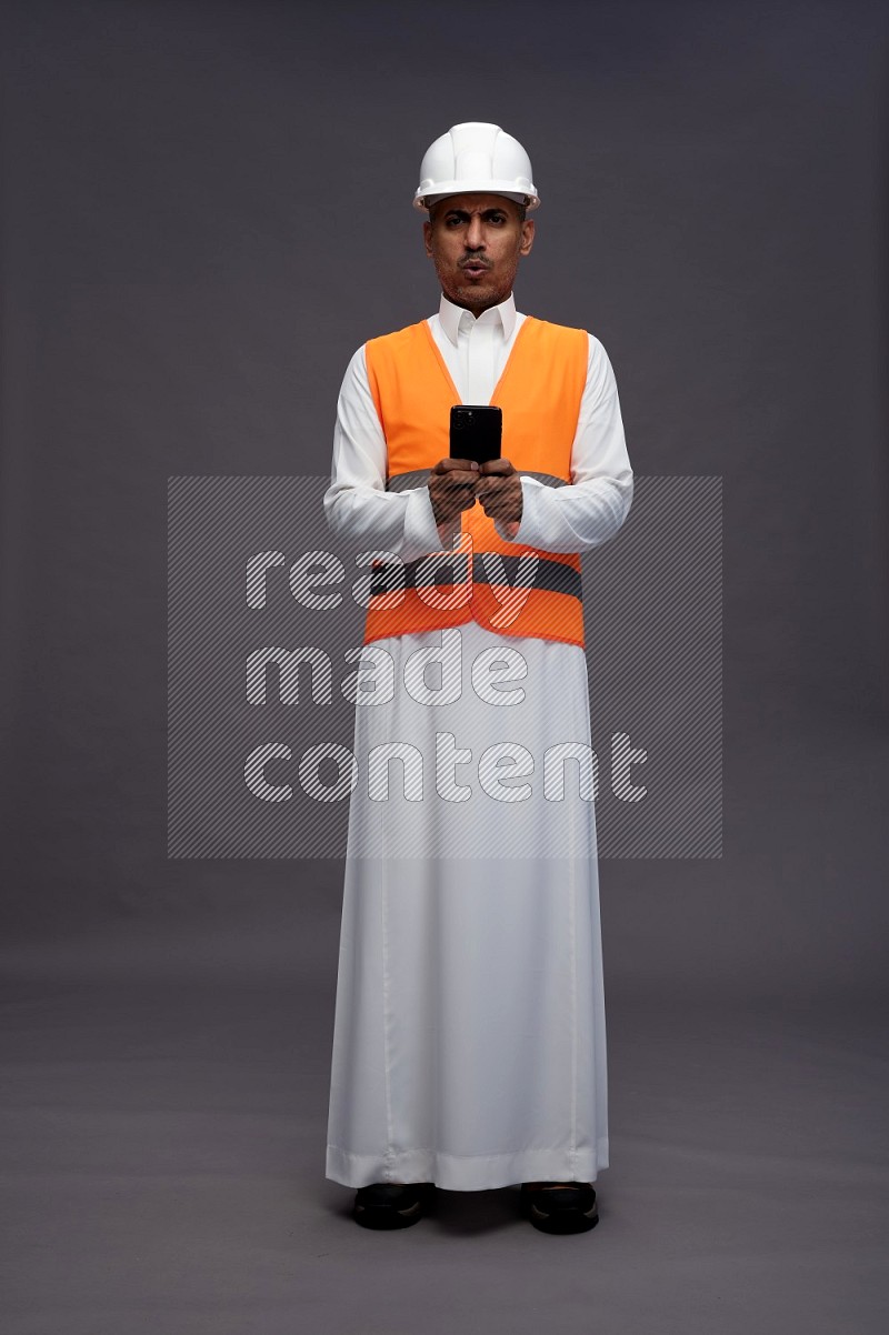 Saudi man wearing thob with engineer vest standing texting on phone on gray background