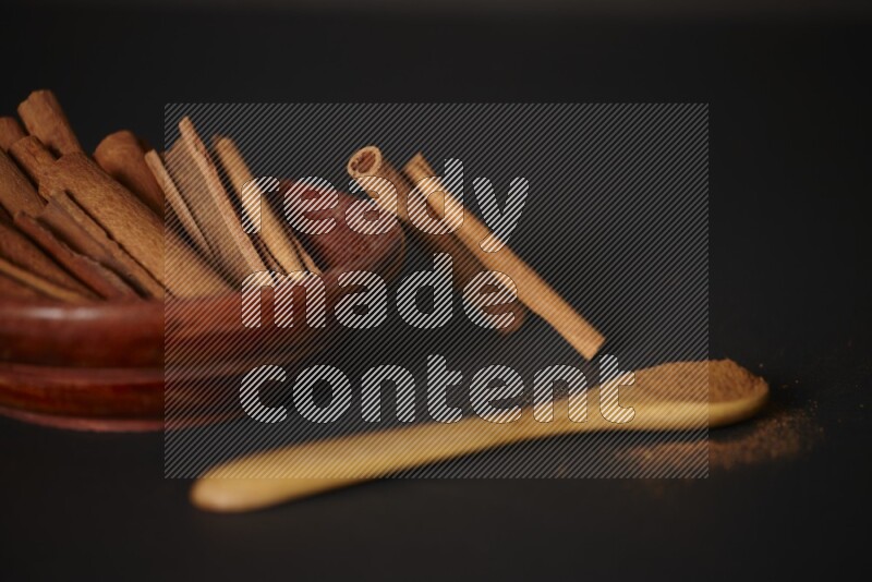 Cinnamon sticks in wooden bowl and cinnamon powder in a wooden spoon on black background