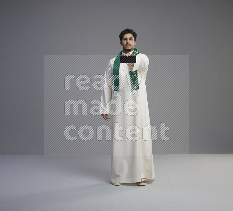 A Saudi man standing wearing thob and Saudi flag scarf showing phone to camera on gray background