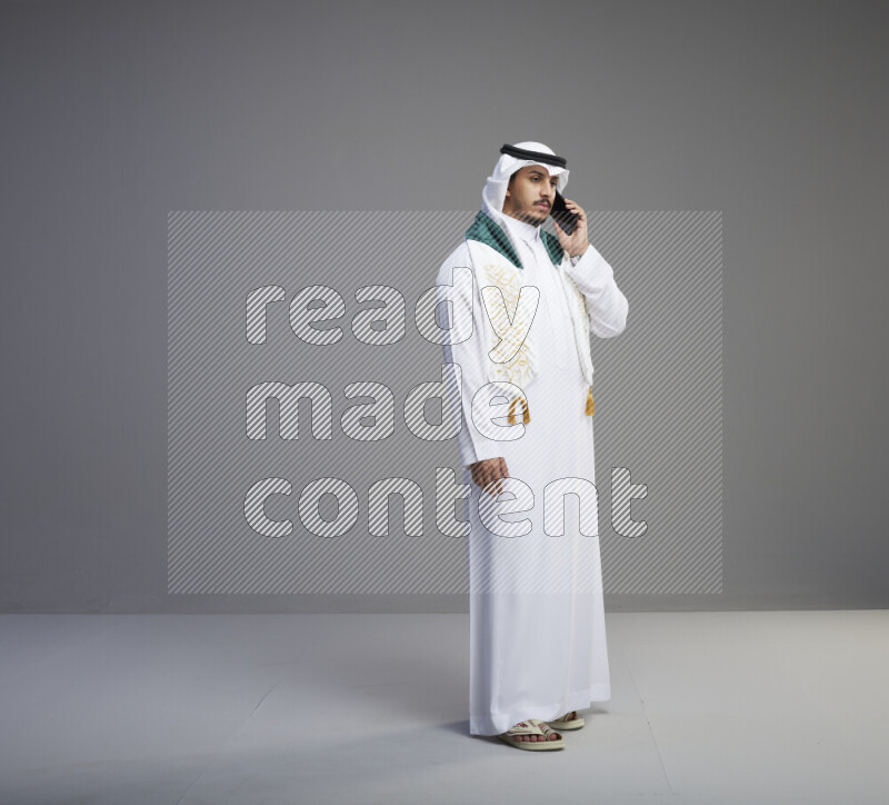 A saudi man standing wearing thob and white shomag with flag scarf on his neck talking on phone on gray background