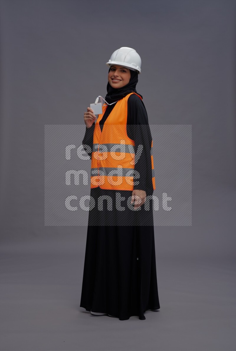 Saudi woman wearing Abaya with engineer vest with neck strap employee badge standing interacting with the camera on gray background